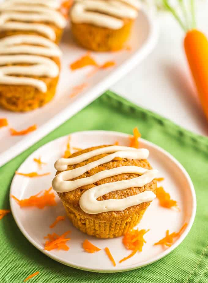 Close up side angle shot of a whole wheat carrot cake muffin with cream cheese glaze and shredded carrots on a white plate with a green napkin underneath