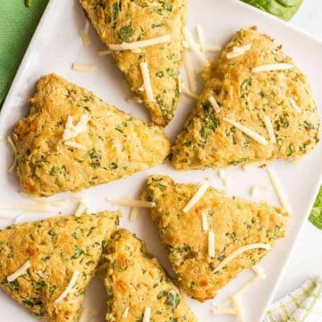 Six whole wheat spinach cheddar scones arranged on a white serving tray with a green towel nearby and a sprinkling of spinach leaves and shredded cheddar cheese.