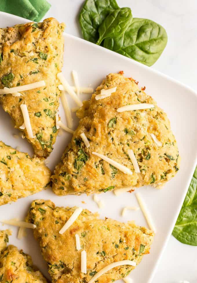 Close up of one whole wheat spinach cheddar scone on a white serving tray with a green towel nearby and a sprinkling of spinach leaves and shredded cheddar cheese