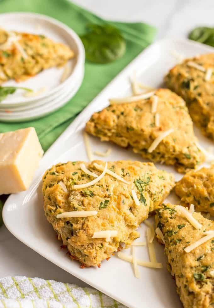 Side angle of whole wheat spinach cheddar scones arranged on a white serving tray with a green towel nearby and a sprinkling of spinach leaves and shredded cheddar cheese and a white serving plate with a scone in the background