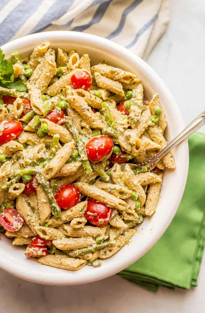 Overhead photo of a large white bowl of creamy pesto pasta with spring vegetables and a serving spoon on the side of the bowl