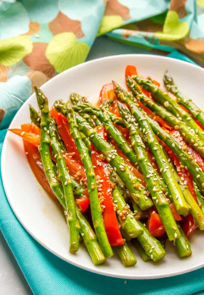 Side angle of sauteed sesame asparagus and red bell peppers on a white plate with colorful napkins in the background