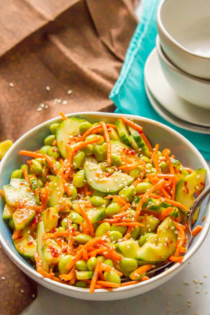 A spoon resting in a blue tinted bowl with a veggie salad of carrots, cucumber and edamame in a soy style dressing with sesame seeds on top.
