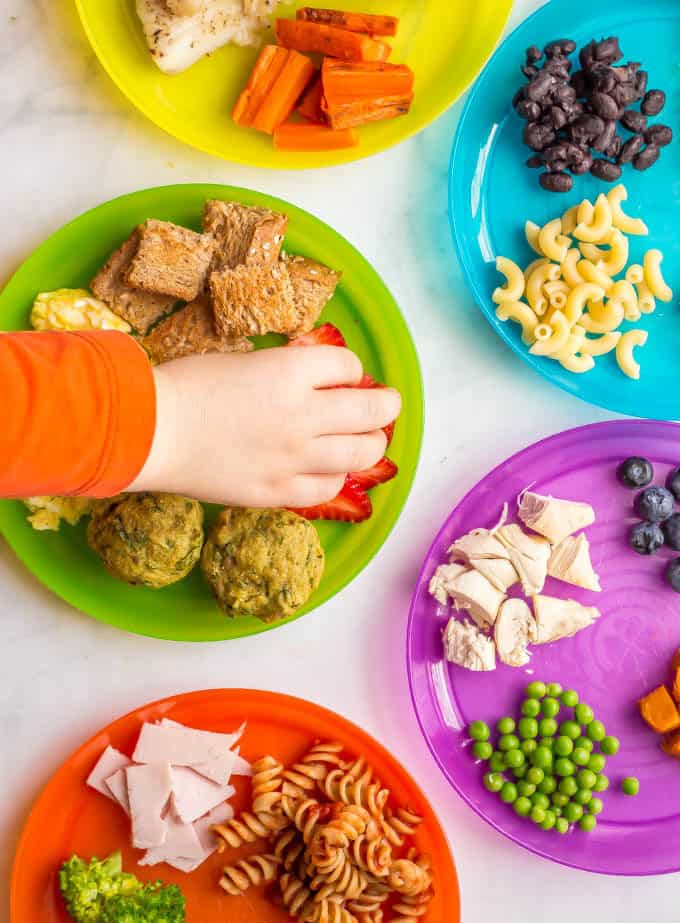 Overhead image of colorful plates with a variety of healthy baby finger foods and toddler finger foods with a small hand reaching in