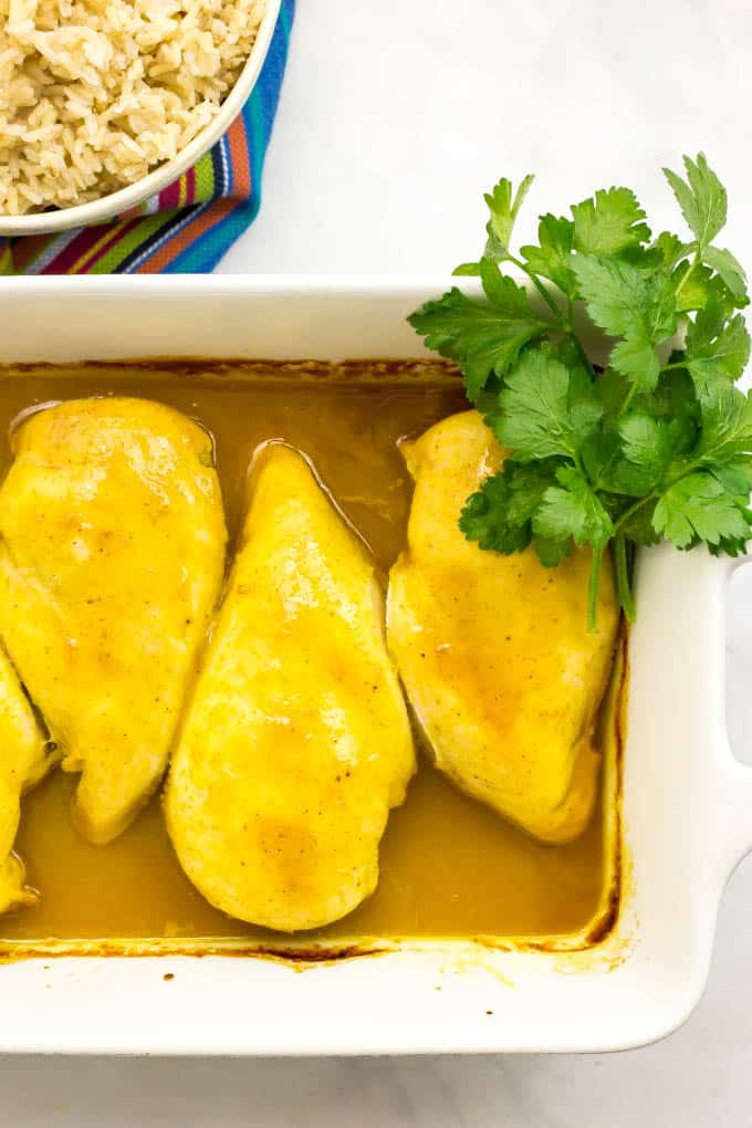 Close up of honey butter chicken in a white casserole dish with a bunch of parsley on the side and a bowl of rice in the background