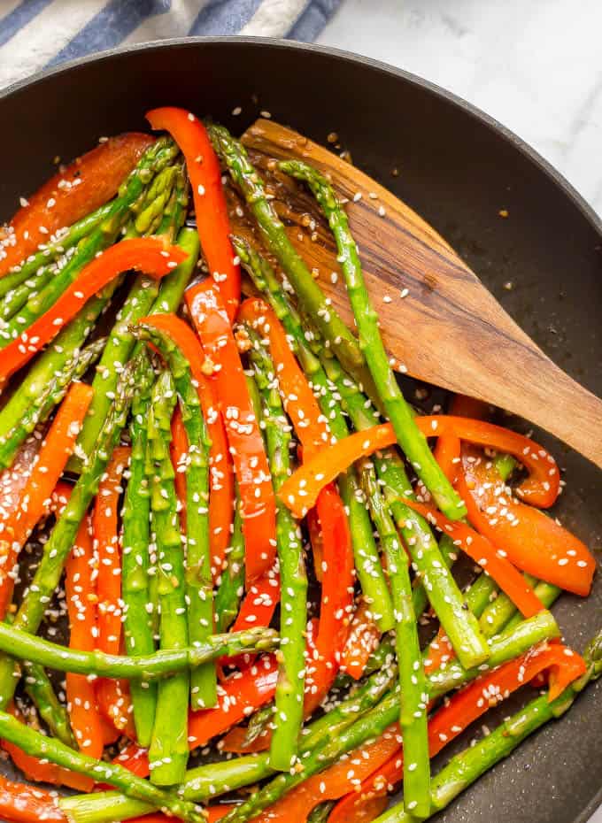 Close- up of sesame asparagus and red peppers in a saute pan with a wooden spoon and a sprinkling of sesame seeds