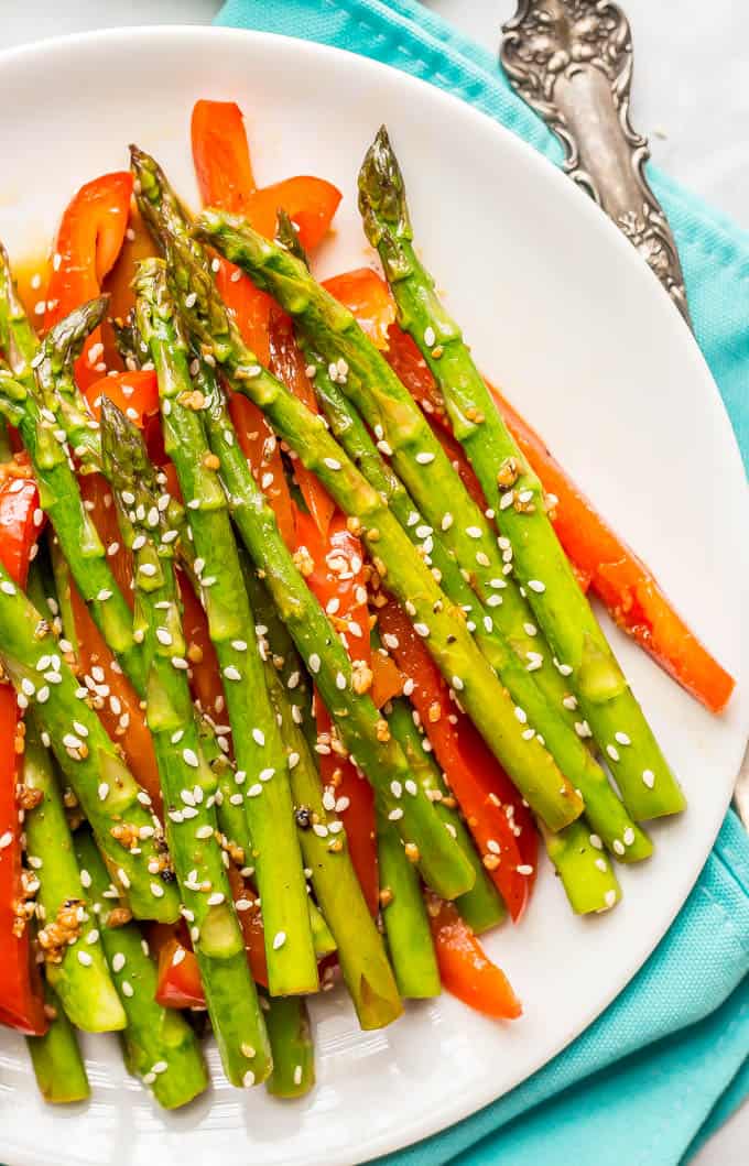 Close-up of sesame asparagus and red peppers on a round white plate with a napkin and serving fork in the background