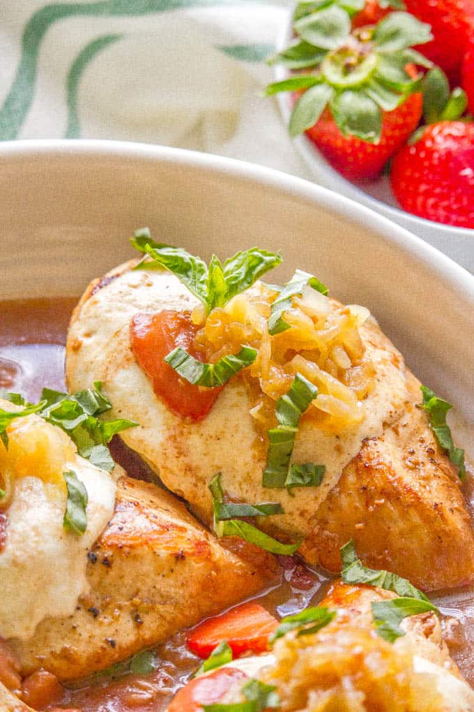 Close up photo of strawberry balsamic chicken with mozzarella in a white dish with a bowl of strawberries in the background