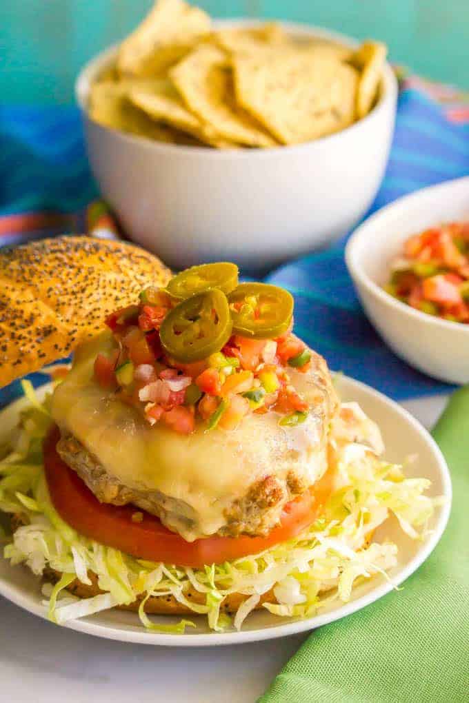A turkey burger served over lettuce and tomato on a bun with melted cheese, pico de gallo and pickled jalapenos on top plus chips in the background.