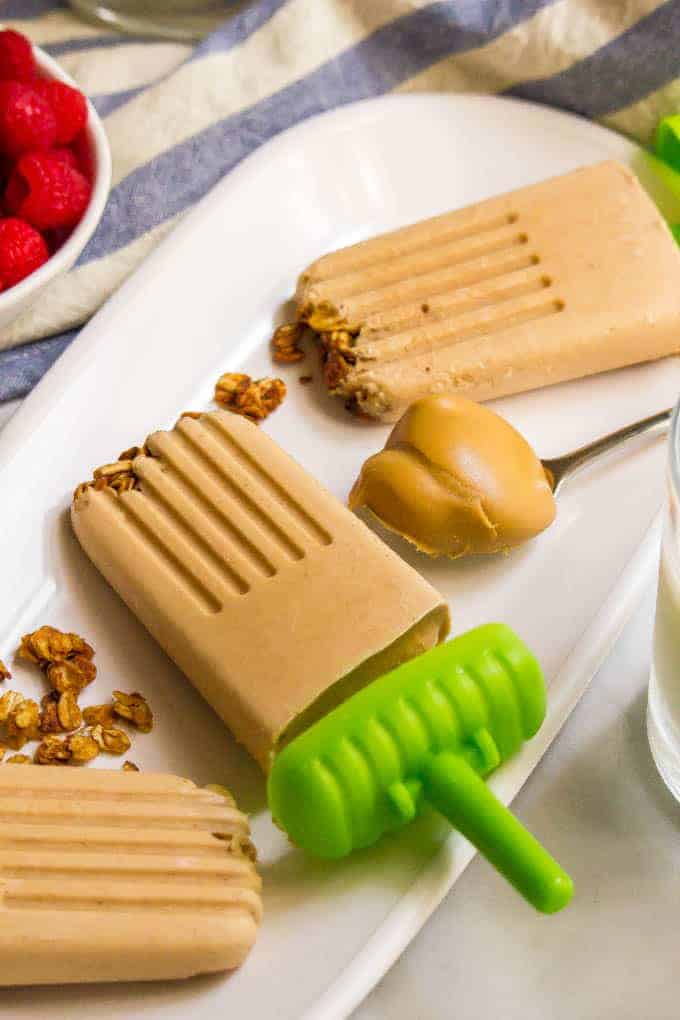 These yummy breakfast popsicles are naturally sweetened and made with just 5 healthy ingredients. The creamy peanut butter and banana flavor is addictive and the crushed cereal or granola brings a fun texture! #popsicles #summereats #kidsbreakfast #frozentreats #healthybreakfast Peanut butter banana breakfast popsicles resting on an oval tray with granola scattered about and a spoonful of peanut butter resting to the side.