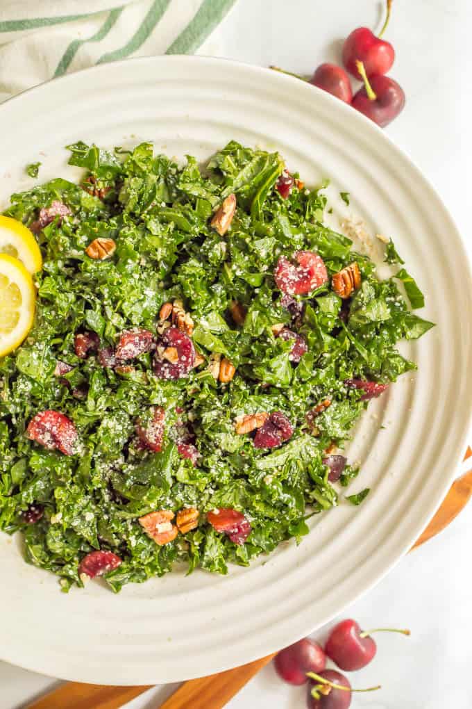 Kale salad in a large white bowl with cherries and pecans mixed in and lemon slices to the side for garnish.