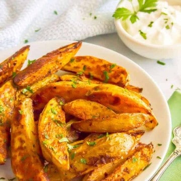 Spicy roasted potato wedges on a white plate with parsley scattered on top with a bowl of sour cream in the background for dipping.