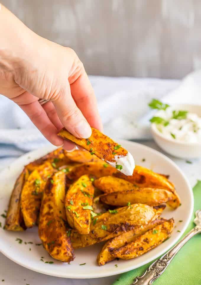 Easy spicy oven roasted potatoes on a white plate with one dipped in sour cream and being held