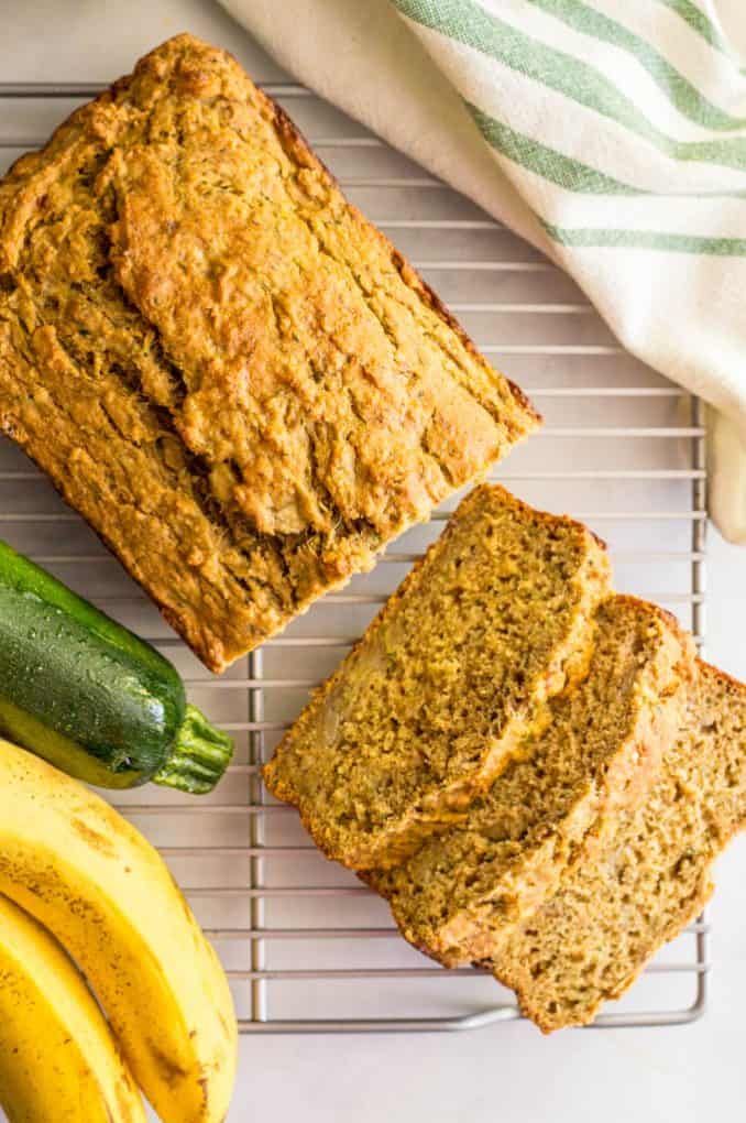 A loaf of zucchini banana bread on a cooling rack with a few pieces sliced alongside a zucchini and banana.
