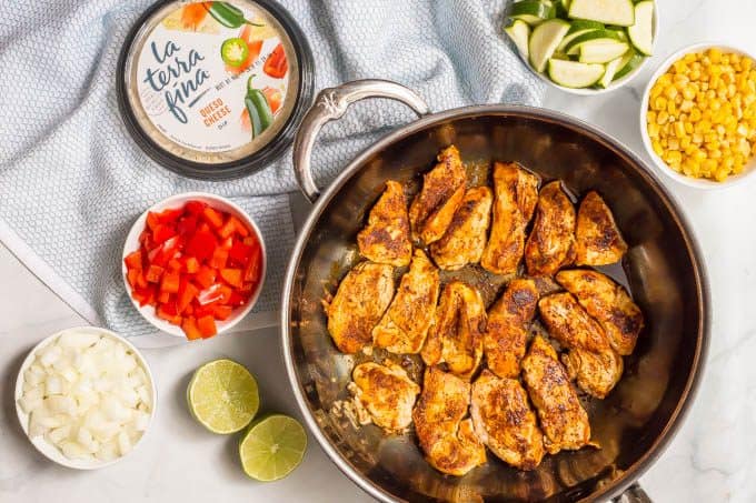 Queso chicken skillet with vegetables ingredients laid out on a marble board with the chicken seared in the pan