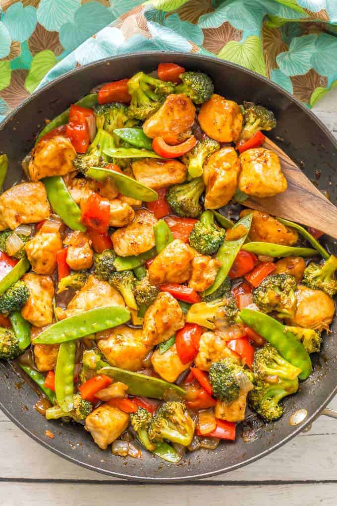 A wooden spatula resting in a large dark skillet full of a homemade sweet and sour chicken with broccoli, peppers and snow peas.
