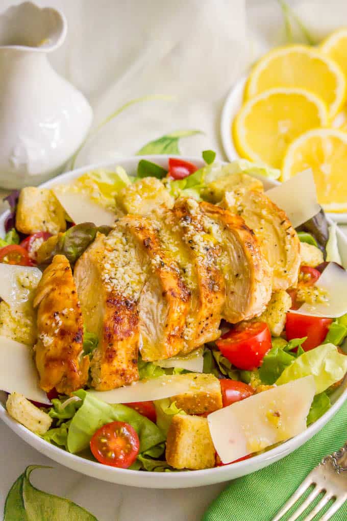 Sliced chicken breast sprinkled with Parmesan cheese and served over a homemade Caesar salad with lemon slices in the background.