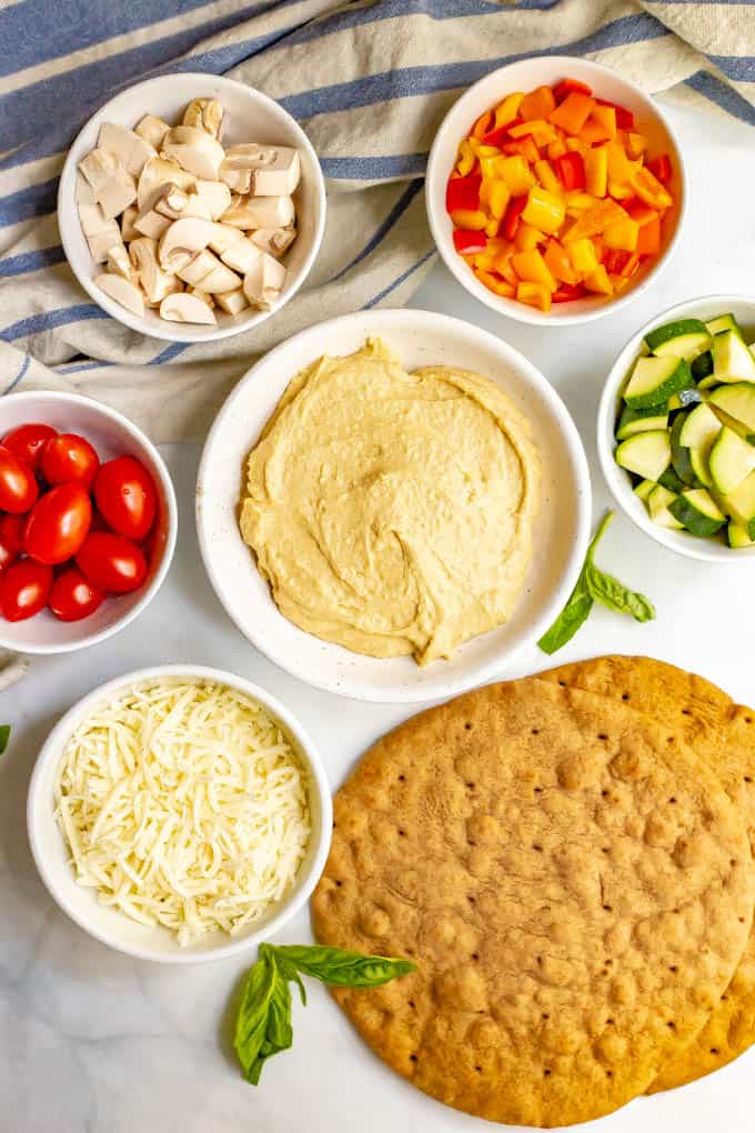 Ingredients for hummus pita pizza with veggies in separate bowls laid out on counter