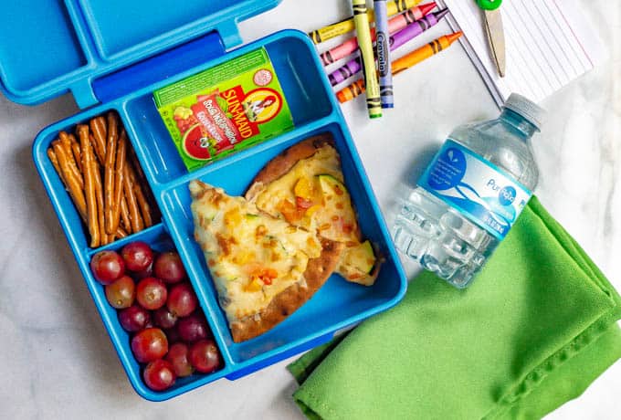 Blue lunchbox with hummus pita pizza with veggies, grapes, pretzels, raisins and a water bottle nearby