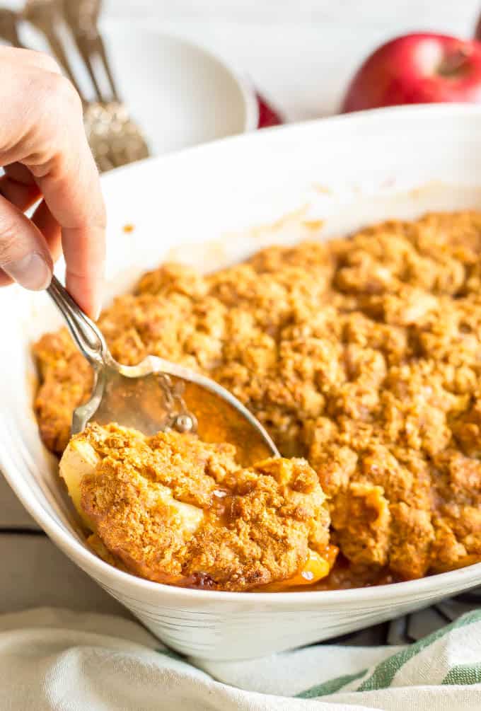 A scoop being taken out of an apple cobbler in a white rectangular baking dish with a silver serving spoon.