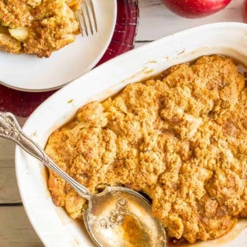A silver serving spoon resting in a white baking dish full of a homemade apple cobbler with a serving on a plate nearby.