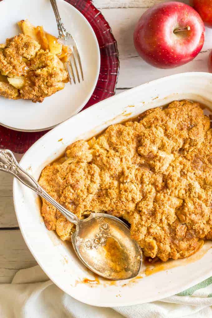 This healthy apple cobbler is full of apple and cinnamon flavors and goes perfect with ice cream for a delicious dessert. It’s naturally sweetened and made with whole grains for a healthier sweet treat that will surely satisfy. #apples #cobbler #dessert #baking A silver serving spoon resting in a white baking dish full of a homemade apple cobbler with a serving on a plate nearby.