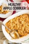 A silver serving spoon resting in a white baking dish full of a homemade apple cobbler with a serving scooped onto a plate nearby.