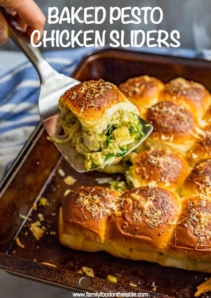 Pesto chicken sliders in a dark baking tray with one being held up by a spatula.
