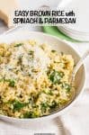 A white bowl of brown rice mixed with spinach and Parmesan and a spoon resting to the side alongside a hunk of fresh Parmesan and a stack of small serving plates.