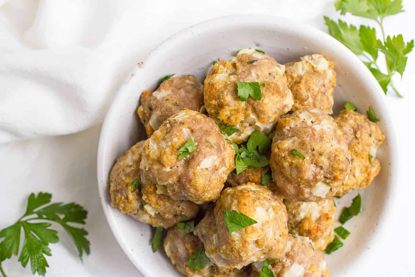 A pile of baked meatballs in a white bowl with fresh parsley sprinkled over top.