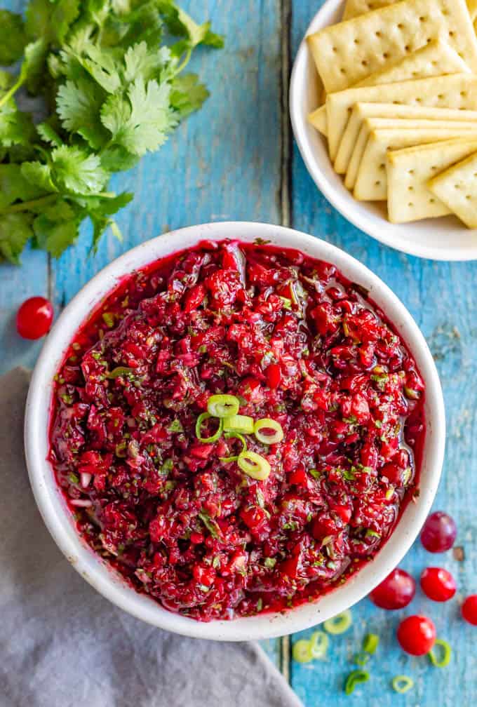 Overhead of fresh cranberry salsa in a white bowl with crackers and cilantro nearby