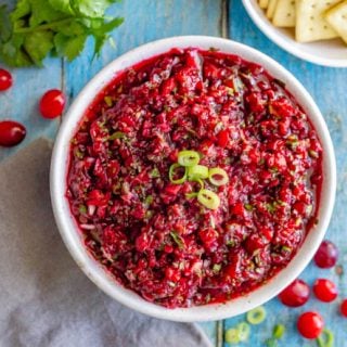 Overhead of fresh cranberry salsa in a white bowl on a turquoise table with cranberries and green onions sprinkled around