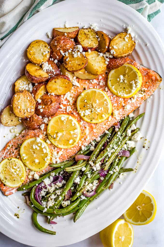 A serving platter with a large slab of salmon topped with lemon slices alongside roasted potatoes and green beans, with feta cheese sprinkled on top.