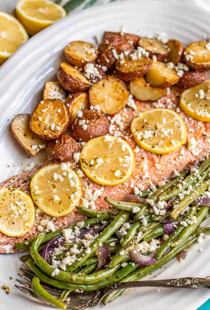 A large piece of roasted salmon in a serving platter with lemon slices on top and roasted potatoes and green beans to the sides.
