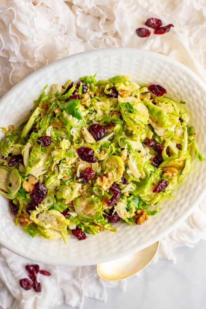 Shredded Brussels sprouts with dried cranberries and walnuts in a white serving dish with a serving spoon nearby