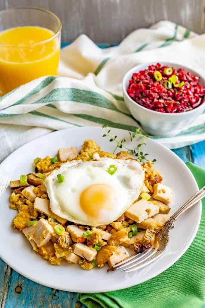 A breakfast hash with Thanksgiving leftover turkey and stuffing and veggies topped with a fried egg and served on a white round plate with a fork.