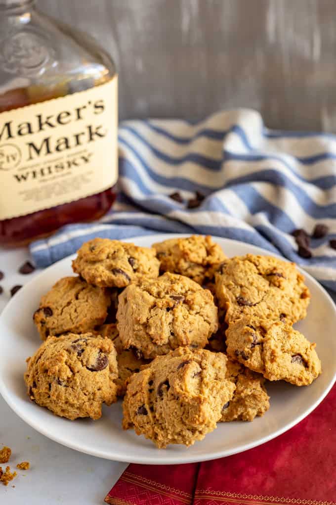 A plate full of bourbon chocolate chip cookies with a bottle of Maker's Mark and a blue striped towel in the background.