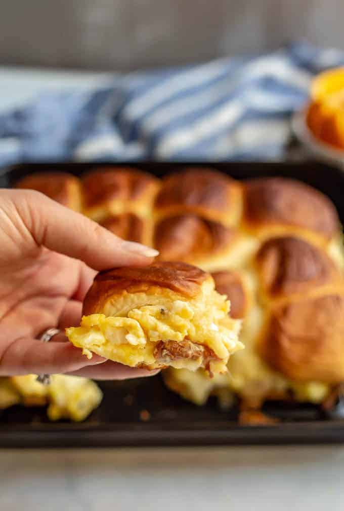 A hand holding up a cheesy scrambled egg and bacon breakfast slider from a tray of sliders.