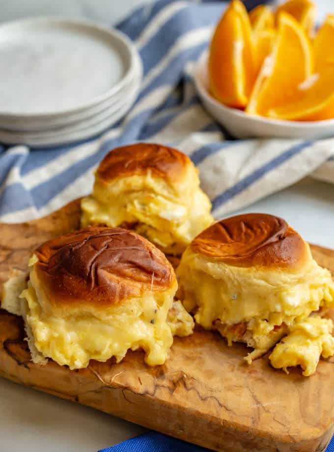 3 cheesy baked breakfast sliders served on a small cutting board with a bowl of oranges in the background