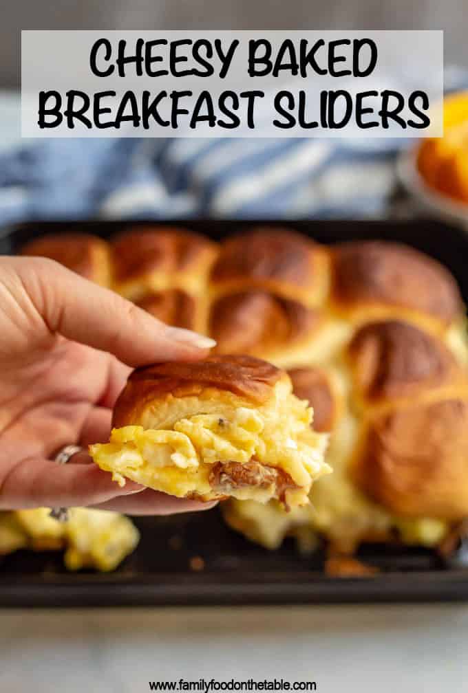 A hand holding up a cheesy egg and bacon breakfast slider from a tray of sliders.