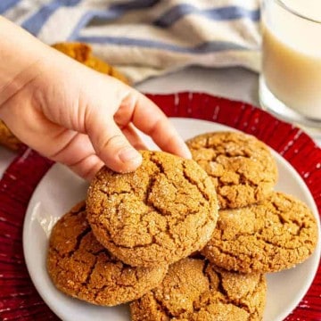 A hand picking up a molasses cookie from a white plate with a glass of milk in the background.
