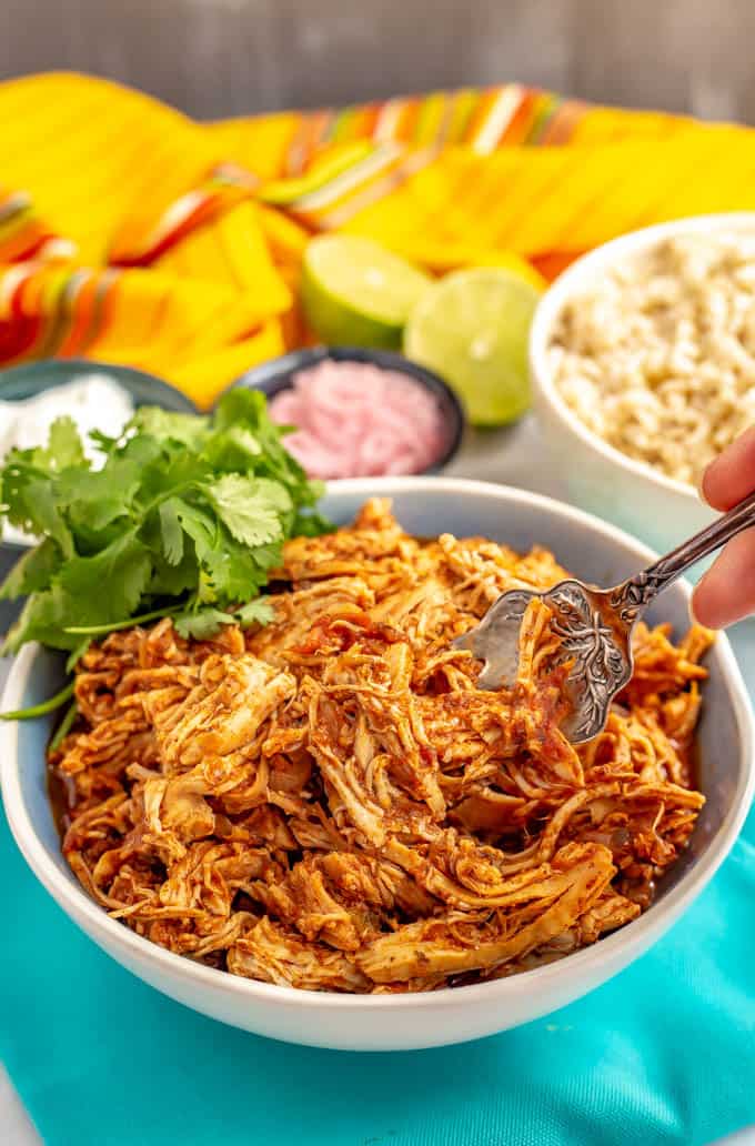 A forkful of slow cooker Mexican shredded chicken being taken from a bowl with sides and toppings in the background