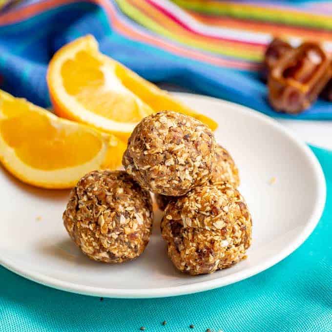 Four peanut butter energy balls on a small white plate with orange slices with some dates in the background.