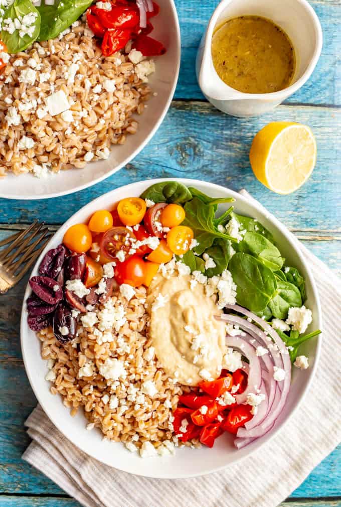 Two vegetarian grain bowls with farro, greens, tomatoes, olives, red onion and hummus with feta cheese over top.