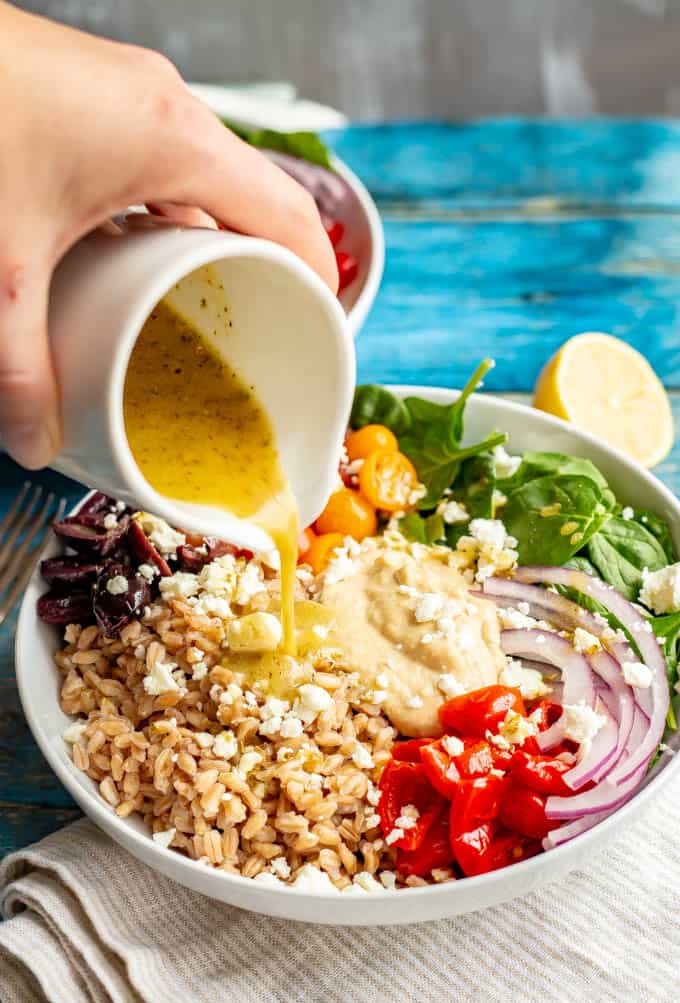A hand pouring a vinaigrette over a Greek style grain bowl of farro with veggies, olives, hummus and feta cheese.