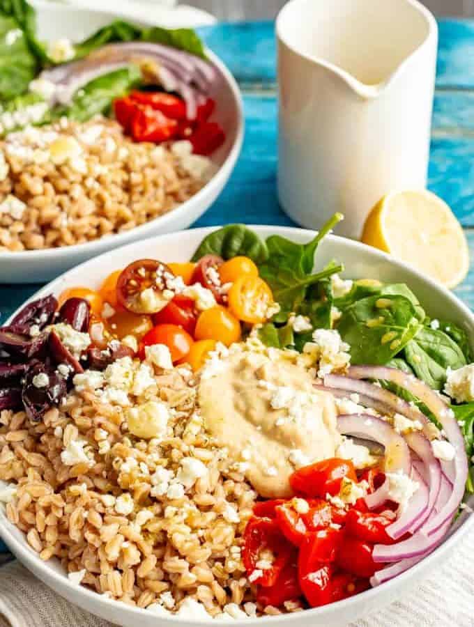 Two Greek style vegetarian grain bowls with farro, greens, tomatoes, olives, red onion and hummus with feta cheese over top.