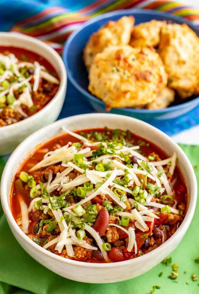 Bowl of Super Bowl chili with toppings and a cheddar biscuits in a separate bowl nearby