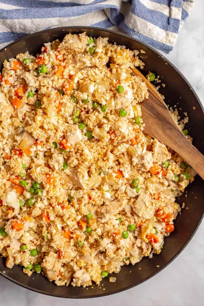 A wooden spatula resting in a large dark skillet filled with a cauliflower fried rice mixture with chicken, peas and carrots.