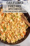 A wooden spatula resting in a dark skillet of cauliflower fried rice with chicken and veggies.