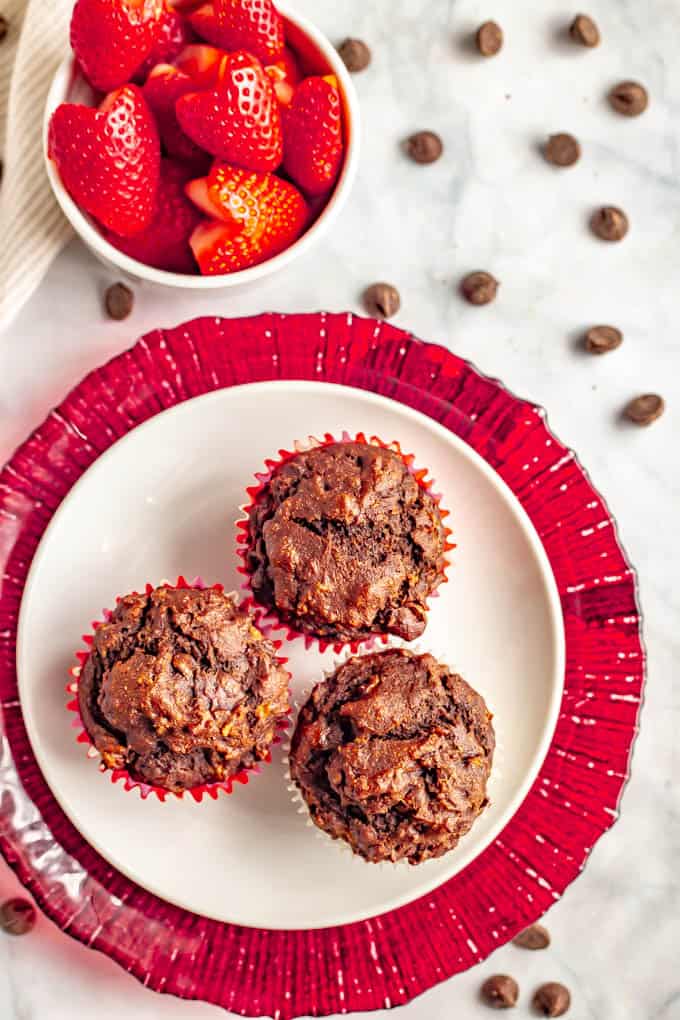 3 double chocolate banana muffins on a white plate with chocolate chips scattered and a bowl of strawberries